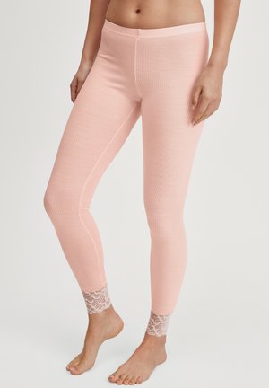 Frau in rosa gerippten Leggings mit Spitzenbesatz an den Knöcheln, die barfuß vor einem schlichten Hintergrund steht.
