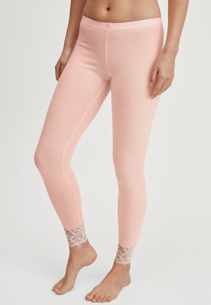 Vrouw in roze geribbelde leggings met kant aan de enkels, staande op blote voeten tegen een effen achtergrond.