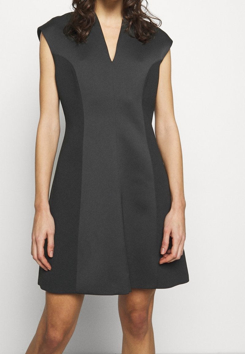 Ärmlös svart klänning med V-ringning och subtila sömnadsdetaljer, bärs av en person med vågigt hår som står mot en vit bakgrund.