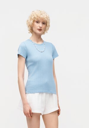 Γυναίκα με κοντά ξανθά σγουρά μαλλιά που φοράει ένα ανοιχτό μπλε ριμπ μπλουζάκι με κοντά μανίκια, λευκά σορτς και ένα κολιέ με χάντρες σε λευκό φόντο.