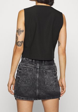 Person trägt ein schwarzes ärmelloses Top und einen ausgebleichten schwarzen Denim-Minirock, zeigt Tätowierungen einer Biene, einer Schlange und eines kreisförmigen Designs auf den Armen.