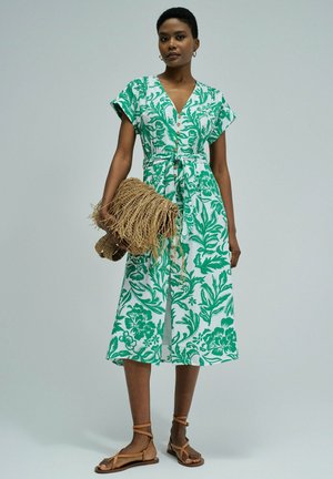Mujer con vestido midi de estampado floral verde y blanco, sujetando un bolso de mano de paja con flecos y llevando sandalias planas color marrón claro con tiras.