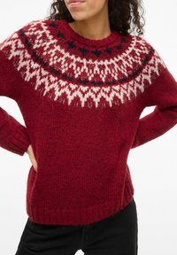 Pull en tricot rouge avec un col rond, présentant un empiècement à motifs blancs et noirs. Texture en tricot, manches longues et ourlet côtelé.
