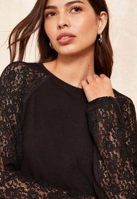 Top nero con un corpo in tessuto liscio e intricate maniche lunghe in pizzo con motivi floreali. Il collo è rotondo e la vestibilità è rilassata.
