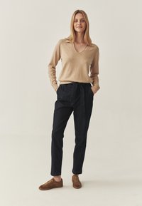 Beige V-Ausschnitt-Pullover kombiniert mit dunklen Jeanshosen mit gebundenem Bund. Das Model trägt braune Wildledersneaker. Neutraler Hintergrund.