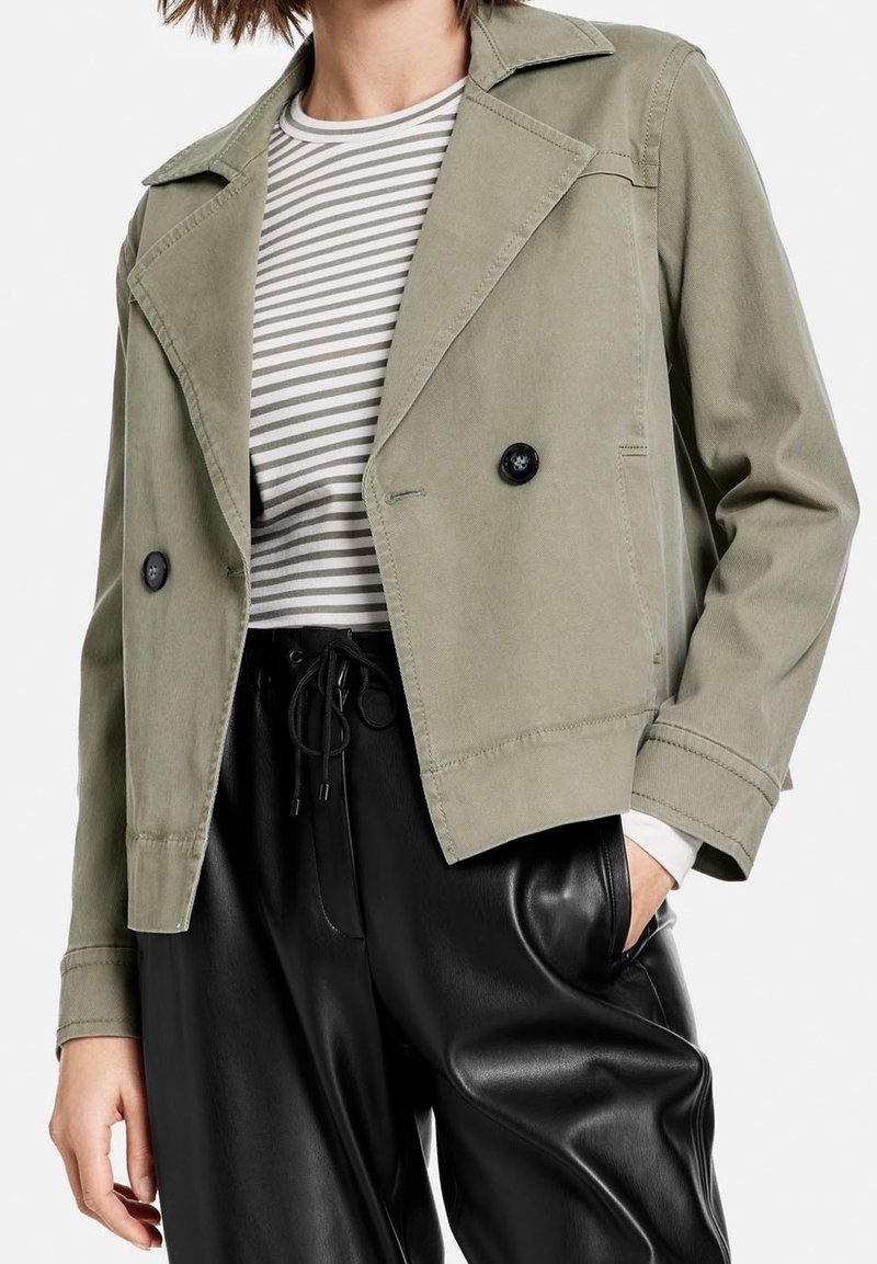 Krótkia kurtka oliwkowo-zielona z kołnierzem z wycięciem, podwójnym czarnym zapięciem na guziki i subtelnymi detalami ściegów, zestawiona z koszulą w paski i czarnymi spodniami.
