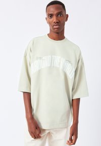 Light green oversized t-shirt made of soft material, featuring bold white text "AROUND THE WORLD" and minimal branding details.
