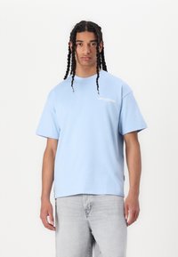 Lys blå bomull T-skjorte med korte ermer, rund halslinje, og en frontlogo med teksten "LES CLASSIQUES." Kombinert med lysegrå jeans.