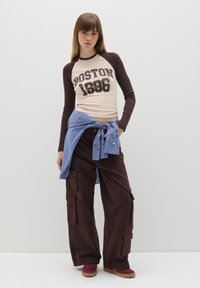 Braunes Langarmshirt mit dem Aufdruck "BOSTON 1996" auf der Vorderseite, kombiniert mit braunen Cargo-Hosen und einem blau karierten Hemd, das um die Taille gebunden ist.