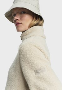 Sudadera de felpa color crema con cuello alto y superficie texturizada, que presenta un parche de logo en la manga izquierda y un sombrero tipo bucket de color claro.