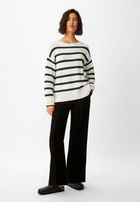 Gestreifter Pullover in Creme und Schwarz, gestrickte Textur, weiter Halsausschnitt, kombiniert mit lockeren schwarzen Hosen und schwarzen Slippers.