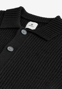Cardigan noir texturé avec un design à côtes, doté d'un col plat, de deux boutons gris et d'une étiquette de marque à l'intérieur du col.
