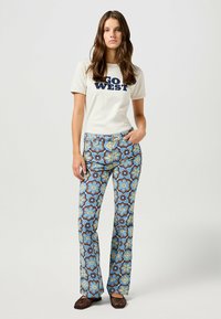 Jeans acampanados con estampado floral en azul, marrón y amarillo, combinados con una camiseta crema con texto en azul marino. Zapatos de esparto marrones completan el look.