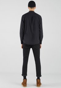 Camicia nera con bottoni e maniche lunghe, jeans scuri e stivaletti marroni alla caviglia. La camicia presenta un yoke posteriore e polsini arrotolati.