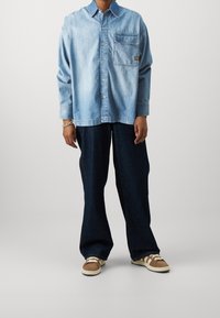Jasnoniebieska koszula jeansowa na guziki z długimi rękawami i kieszenią na piersi, zestawiona z ciemnoniebieskimi dżinsami o szerokich nogawkach i brązowymi trampkami.