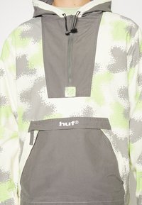 Pullover com capuz em padrão de camuflagem cinza e verde, com um bolso frontal e detalhe de logotipo. O material parece leve e durável.