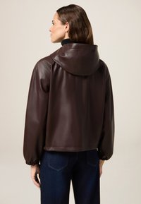 Giubbotto in pelle marrone con cappuccio, dal taglio comodo e polsini elastici. Indossato sopra un jeans scuro, che mette in risalto una texture morbida.