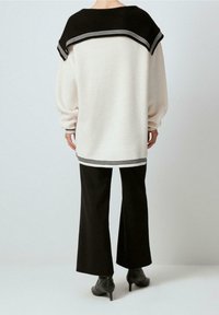 Übergroßer cremefarbener Pullover mit schwarzen Schulterakzenten und einem gestreiften Saum, kombiniert mit schwarzen Hosen mit weitem Bein und Stiefeln mit Absatz. Glatte Textur.