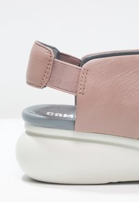 Primo piano di un sandalo in pelle rosa con cinturino elastico sul retro, soletta imbottita grigia e suola spessa e morbida bianca.