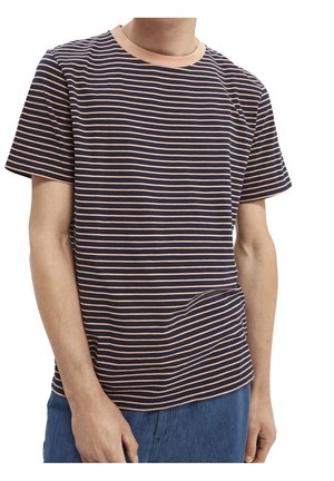 Person iført en kortærmet marineblå t-shirt med tynde lyserøde vandrette striber og blå bukser, stående mod en hvid baggrund.