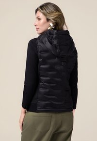 Schwarze Steppweste mit Kapuze, mit gestepptem Muster und glänzender Oberfläche. Getragen über einem schwarzen Oberteil und kombiniert mit olivgrünen Hosen.