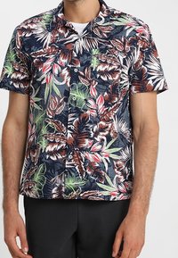 Muž v košili s krátkým rukávem, pokryté vícebarevným tropickým listovým vzorem, a černými kalhotami na jednobarevném pozadí.