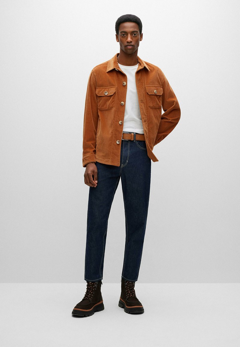 Kordjacke in verbranntem Orange mit zwei Brusttaschen, weißes T-Shirt, blaue Jeans und braune schnürbare Stiefel mit einer robusten Sohle.
