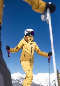 Geel en bruin skikostuum met ritszakken, zwarte handschoenen en skistokken. Witte helm met vizier en spiegeltjesgoggles. Sneeuwachtige bergen op de achtergrond.