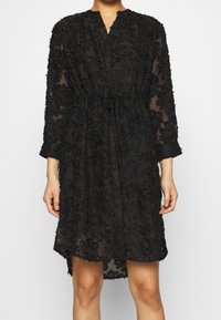 Vestido negro hecho de tejido transparente, con bordados florales, cintura con cordón y mangas de tres cuartos, con un dobladillo ligeramente asimétrico.