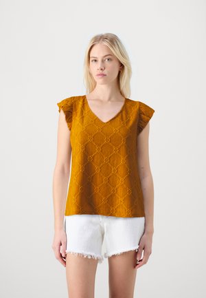 Blouse jaune moutarde avec un design texturé à motifs de diamant, manches courtes à volants et décolleté en V. Associée à un short blanc effiloché.