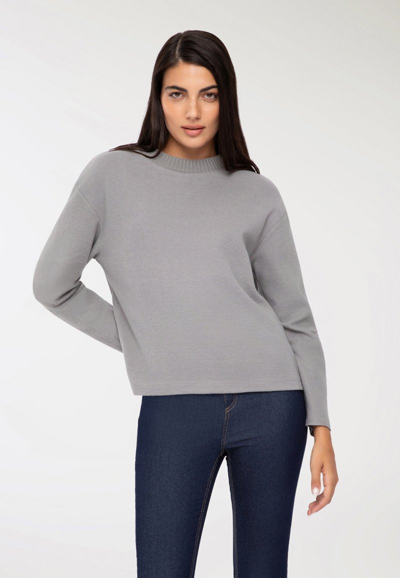 Szary sweter z okrągłym dekoltem, długimi rękawami i luźnym krojem, zestawiony z ciemnoniebieskimi dżinsami. Materiał wydaje się miękki i gładki.