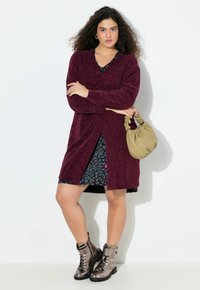 Burgundy texturerad kofta med långa ärmar, bärs över en svart klänning med leopardmönster. Modellen håller en ljusbeige handväska.