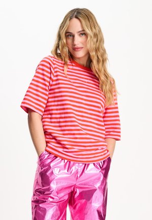 Vrouw met lang golvend blond haar draagt een roze en rood gestreept hemd en glanzende roze broek, staand met handen in de zakken.