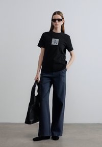 Μαύρο βαμβακερό T-shirt με ασημένιο λογότυπο, συνδυασμένο με ψηλόμεση, φαρδιά παντελόνα σε ναυτικό μπλε, και μαύρη τσάντα χειρός. Το μοντέλο φοράει γυαλιά ηλίου.