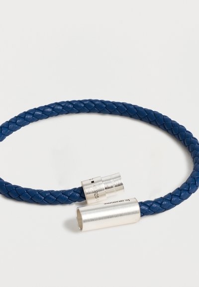 Bracelet en cuir tressé bleu avec un fermoir magnétique cylindrique argenté partiellement ouvert sur fond blanc.