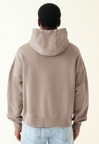 Sudadera amplia color topo con una gran capucha, puños y dobladillo de canalé, y una textura suave. La lleva puesta con jeans de color azul claro.