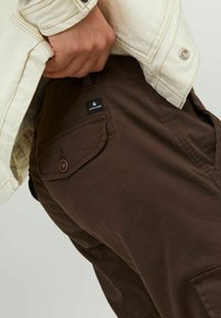 Pantaloni cargo marroni con dettagli di tasche laterali, caratterizzati da un bottone decorativo e un'etichetta con marchio. Il tessuto sembra resistente e dalla texture liscia.