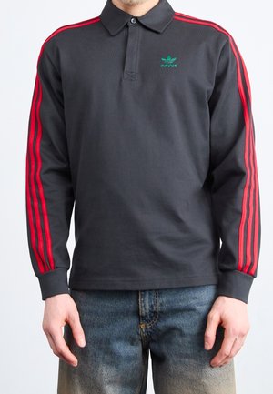 Polo à manches longues noir avec trois bandes rouges sur les manches et logo Adidas vert sur la poitrine, porté avec un jean bleu délavé.