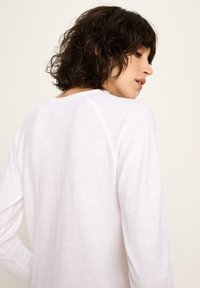 Frau mit kurzen, gewellten schwarzen Haaren, die seitlich schaut, trägt ein einfaches weißes Langarmshirt vor einem beigen Hintergrund.