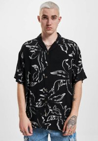 Schwarzes Kurzarmhemd mit weißem botanischem Muster, Knopfleiste, lockerer Schnitt und leichtem Stoff. Ausgebleichte Jeans.