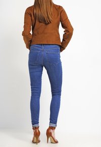 Veste en daim marron, jean skinny bleu, revers léger aux chevilles et chaussures à talons hauts marron. Texture lisse et design ajusté.