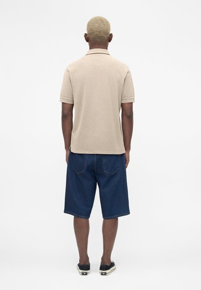 Άνδρας με κοντά ξανθά μαλλιά στέκεται με την πλάτη προς την κάμερα, φοράει ένα μπεζ πόλο με κοντά μανίκια, μπλε τζιν σορτς και μαύρα αθλητικά παπούτσια.