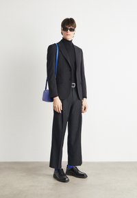 Zwarte suit met een slim-fit ontwerp, gecombineerd met een zwarte coltrui. Zwarte schoenen, blauwe sokken en een kleine blauwe tas. Accessoires omvatten een zonnebril.