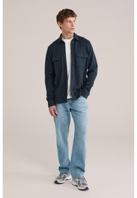 Muž v tmavomodrej bundě na gombíky cez bielu košeľu, svetlomodré džínsy a sivé tenisky, stojaci na pozadí jednoliatej béžovej steny.