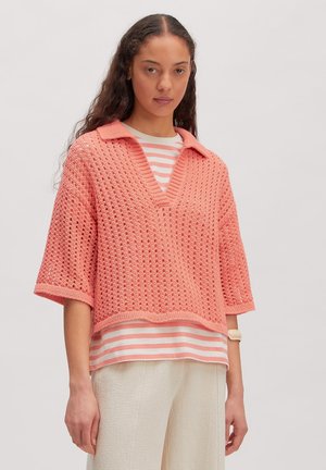 Jonge vrouw draagt een koraalgekleurde gehaakte kortemouwen trui over een wit en koraal gestreept overhemd met crèmekleurige broek.