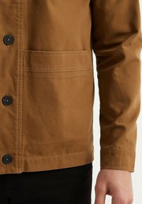 Veste marron en tissu texturé, avec une fermeture à boutons, une poche poitrine et une coupe droite. Les poignets sont boutonnés avec une finition soignée.