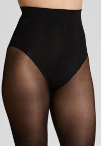 Hoge taille zwarte shapewear onderbroek gedragen over sheer zwarte panty, gefotografeerd van de taille tot halverwege de dij tegen een effen achtergrond.