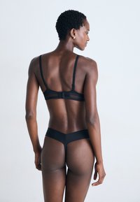 Femme aux cheveux courts et bouclés portant un soutien-gorge noir et un string, se tenant contre un fond clair, de profil avec le dos visible.