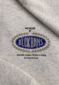 Lichtgrijze stof met een iets gestructureerd oppervlak. Bevat een blauw ovale logo met gouden accenten en de tekst "AANGEDREVEN DOOR BLOCKBNS."