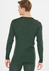 Man met lichtbruin haar, gekleed in een donkergroen lange mouw top en bijpassende broek, van achter gezien tegen een witte achtergrond.
